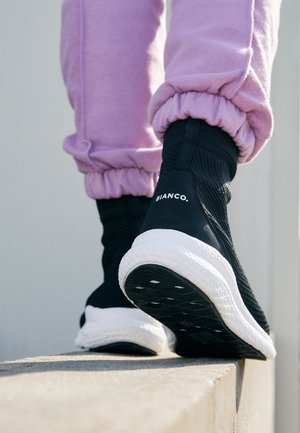 Schwarz-weiße Bianco-Sneaker, getragen mit pinken Jogginghosen, auf einer Betonkante laufend, von hinten und unten gezeigt.