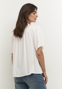 White short-sleeve blouse with a relaxed fit, featuring gathered detail at the shoulders, smooth texture, and a curved hem, paired with blue jeans.