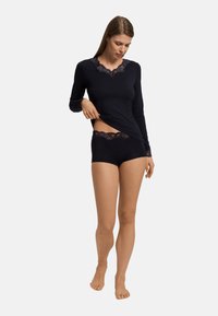 Svart långärmad skjorta och matchande shorts i trosa-stil med spetsdetaljer. Mjuk, elastisk tyg; figurnära design. Modellen justerar skjortan medan hon står barfota.