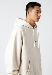 Felpa con cappuccio beige con coulisse, caratterizzata da una tasca a marsupio e da una scritta nera. Tessuto morbido e vestibilità comoda, con dettagli a coste.