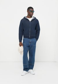 Mörkeblå zip-hettegenser med grå innerside, kombinert med lyseblå jeans og hvite sneakers. Holder en kremfarget caps.