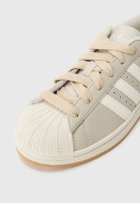 Svetlo béžová teniska s textúrovanou gumovou špičkou, bavlnenými šnúrkami a tromi bielymi bočnými pruhmi. Navrhnutá s hladkým, koži pripomínajúcim zvrškom.