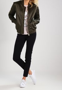 Olivově zelená saténová bomber bunda, szipem vpředu, žebrované manžety, nošena přes světle růžovou košili. Černé slim-fit kalhoty a bílé tenisky.