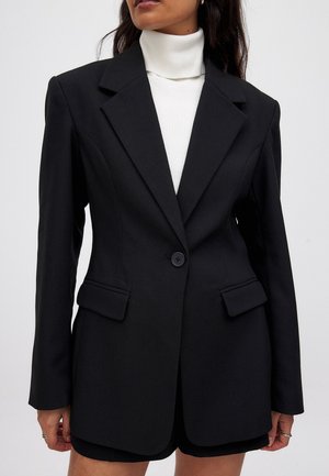 Mujer con blazer negra entallada con solapas de muesca y un solo botón, sobre un jersey de cuello vuelto blanco de canalé.