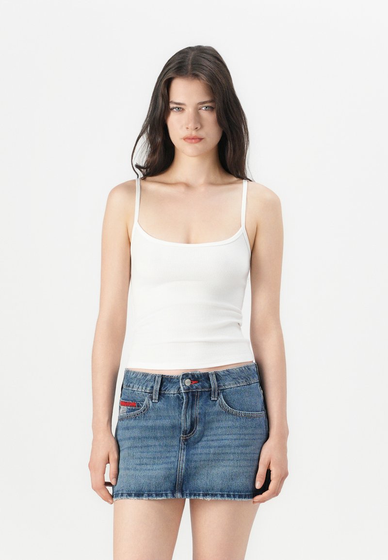 Top de algodón blanco sin mangas con tirantes finos, combinado con una falda mini de mezclilla azul que presenta un dobladillo deshilachado y costuras rojas en los bolsillos.