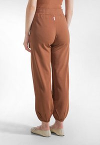 Vista trasera de una persona que lleva pantalones con cintura elástica color óxido y puños fruncidos, junto con zapatos planos beige, de pie sobre un fondo blanco.