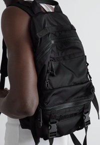 Rucsac negru din nailon cu multiple compartimente cu fermoar, bretele reglabile și accente texturate. Caracteristici hardware cu brand și un design elegant.
