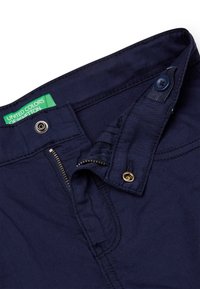Pantalones de algodón azul marino con cierre de cremallera metálica y botón, que presentan una etiqueta de marca verde. Textura suave con sutiles detalles de costura.