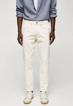 Mann mit hochgekrempeltem langärmligem blauen Hemd, elfenbeinfarbenen Jeans und weißen Sneakers mit beigen Sohlen, der vor einem schlichten hellen Hintergrund steht.