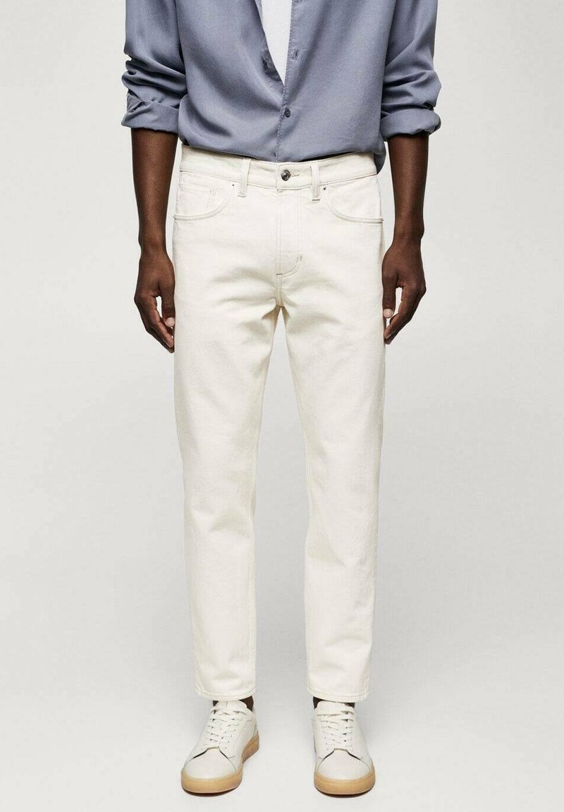 Homme portant une chemise bleue à manches longues retroussées, un jean écru et des baskets blanches avec des semelles beiges, debout sur un fond uni clair.