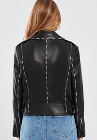 Zwarte leren jas met studs langs de naden, ritsmanchetten en een getailleerd ontwerp. Achteraanzicht dat de gladde textuur toont.