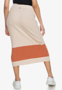 Jupe midi côtelée beige et rouille avec un design bicolore. Elle dispose d'une taille ajustée et d'une texture subtile, portée avec des baskets blanches.