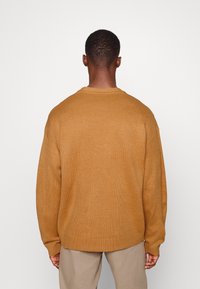 Brun stickad tröja med ribbad struktur, rund halsringning och något oversize passform, kombinerad med beige byxor. Ingen synlig märkning.