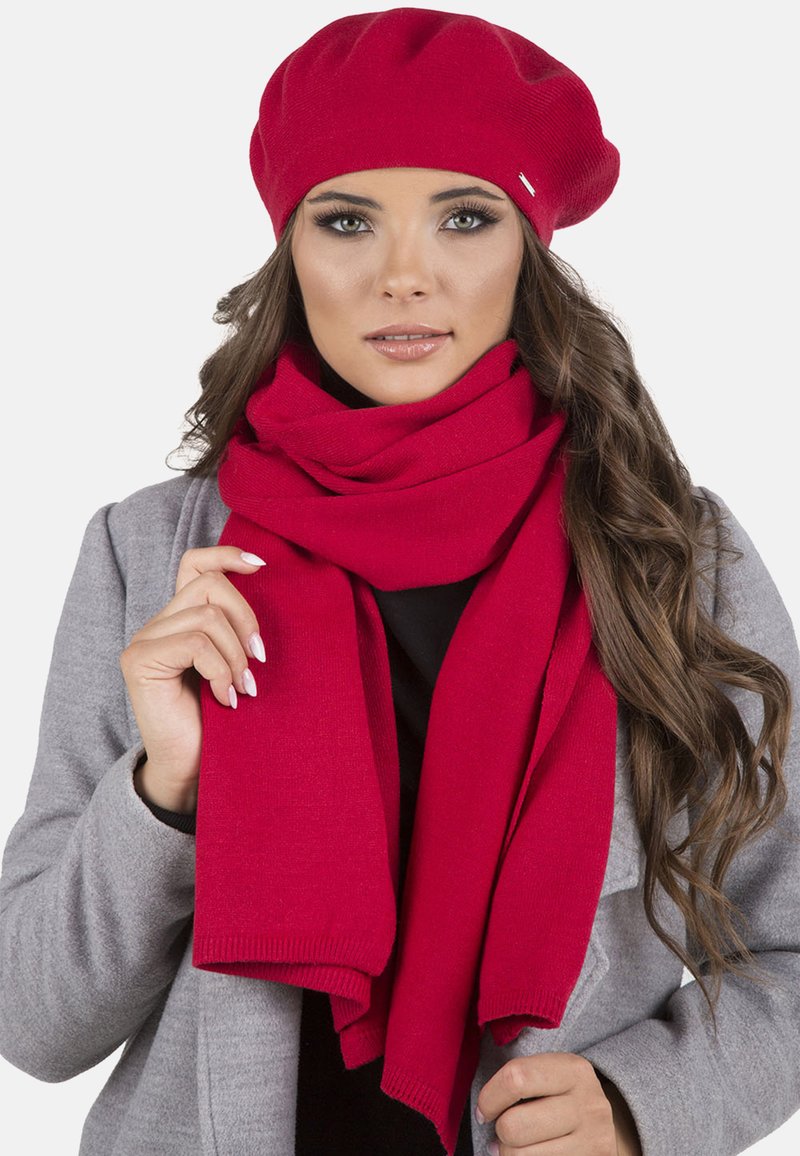 Berretto a maglia rosso e sciarpa coordinata, entrambi senza cuciture e morbidi, in contrasto con un cappotto grigio. La sciarpa cade morbida attorno al collo.