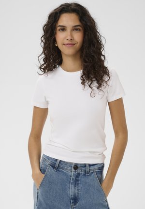 Kobieta z długimi, kręconymi, ciemnymi włosami, ubrana w białą koszulkę z krótkim rękawem i niebieskie dżinsy, stojąca z rękami w kieszeniach na jednolitym tle.