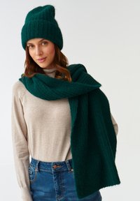 Mørk grøn strikket hue og tørklæde lavet af blødt materiale, båret over en lys beige sweater. Tørklædet hænger løst omkring halsen.