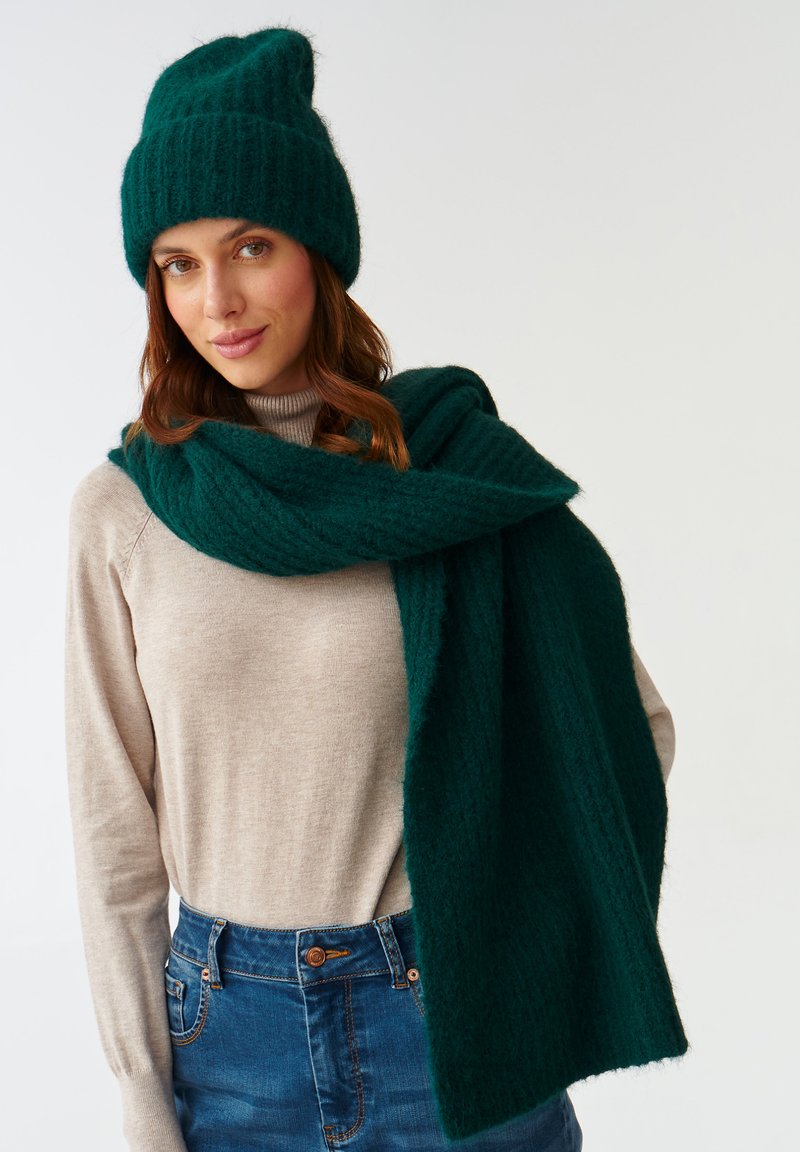 Mørk grøn strikket hue og tørklæde lavet af blødt materiale, båret over en lys beige sweater. Tørklædet hænger løst omkring halsen.