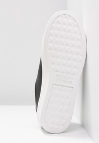 Černá teniska s bílou gumovou podrážkou, která má texturovaný diamantový vzor a vyražené logo na straně podrážky.