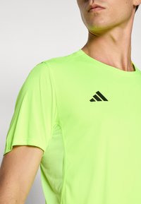 Close-up van een persoon die een felneongroen sportshirt met korte mouwen draagt, met een zwart gestileerd logo op de borst.