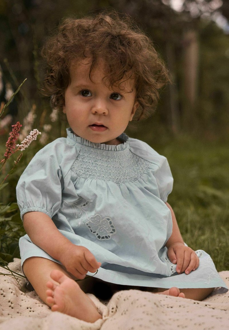 Tout-petit aux cheveux bouclés portant une robe bleu clair, assis pieds nus sur une couverture à l'extérieur, avec de l'herbe et des fleurs sauvages en arrière-plan.