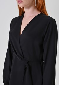 Abito avvolgente nero realizzato in tessuto liscio, con scollo a V profondo, maniche lunghe e un dettaglio in vita con lacci per una silhouette aderente.