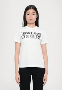 Balta medvilnė T-shirt marškiniai su drąsiu juodu tekstu, kuris sako "VERSACE JEANS COUTURE". Trumpas rankoves ir apvalios kaklo dizainas.