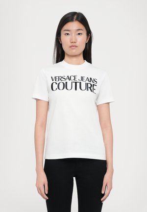 Balta kokvilnas T-krekls ar uzrakstu "VERSACE JEANS COUTURE" treknajā melnajā tekstā. Īsas piedurknes un apaļš kakls.