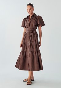 Braunes Baumwollhemdkleid mit kurzen Puffärmel, einem Taillengürtel und einem gestuften Rockdesign, das bis zur Mitte der Wade reicht. Minimalistische Schuhe.