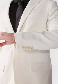 Weißes Leinenanzugjackett mit einer Brusttasche und vier hellen Knöpfen an den Ärmelbündchen. Der Stoff hat eine dezente Textur.