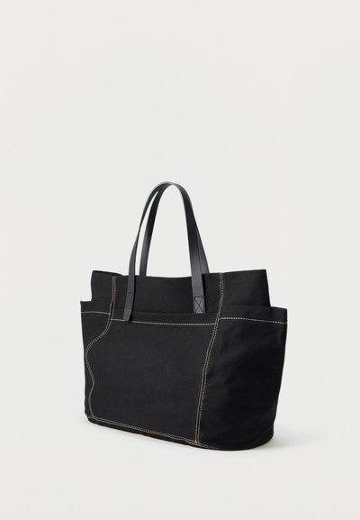 Czarna torba typu tote z płótna z kontrastującym przeszyciem i dwoma czarnymi skórzanymi uchwytami, stojąca pionowo na jednolitym jasnym tle.