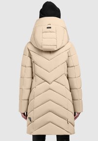 Beige színű puffa kabát, chevron mintával, mely kapucnival, meghosszabbított szilhouette-val és oldalsó hasítékokkal rendelkezik, fekete logó címkével kiegészítve.