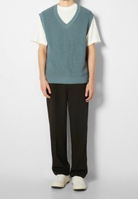 Jasnoniebieska dzianinowa kamizelka z dekoltem w serek, noszona na białej koszulce, w połączeniu z czarnymi spodniami i białymi trampkami. Gładka faktura, swobodny krój.