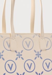 Beige tote-taske lavet af tekstureret materiale med et mønster af blå blomsterdetaljer og cirkulære logoer med et "V." To stærke håndtag.