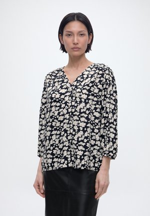 Woman with short dark hair wearing a black and white floral blouse with three-quarter sleeves and a black skirt, facing forward.