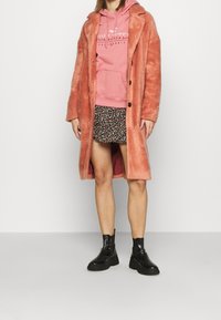 Lång, fluffig rosa kappa med krage, knappstängning och sidofickor, lagd över en blommig kjol och svarta klumpiga ankelstövlar.