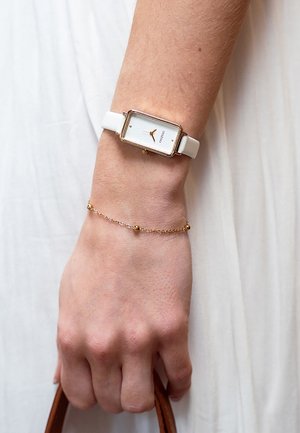Montre blanche avec un boîtier rectangulaire doré, cadran blanc minimaliste, aiguilles dorées, accompagnée d'un délicat bracelet en chaîne dorée.
