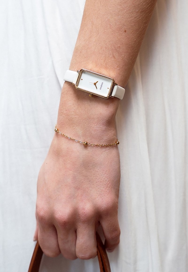 Montre blanche avec un boîtier rectangulaire doré, cadran blanc minimaliste, aiguilles dorées, accompagnée d'un délicat bracelet en chaîne dorée.