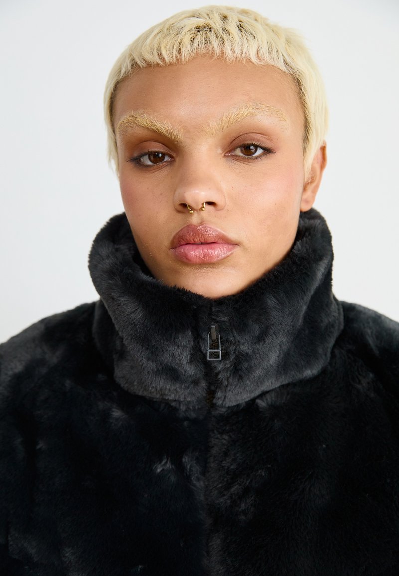Veste en fausse fourrure noire avec un col haut et une fermeture éclair, présentant une texture douce et un léger éclat. Accentuée par un détail de fermeture éclair lisse.