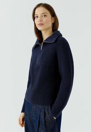 Vrouw die een donkerblauwe, gestructureerde gebreide trui draagt met een halve rits en een brede kraag, gecombineerd met losse donkerblauwe broek.