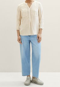 Personne portant un chemisier beige à rayures verticales blanches, un jean large bleu clair et des chaussures à semelle épaisse gris clair.