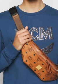 Riñonera de cuero marrón con logotipo en relieve, patrones geométricos, un zipper frontal y correa ajustable a rayas.
