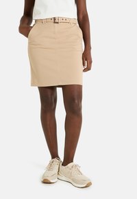 Jupe beige midi à coupe droite, avec deux poches latérales et une ceinture assortie ornée d'accentuations métalliques. Portée avec des baskets beiges.