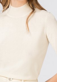 Světle krémový pletený svetr s vysokým límcem a krátkými rukávy, který má hladkou strukturu a přiléhavý silueta.