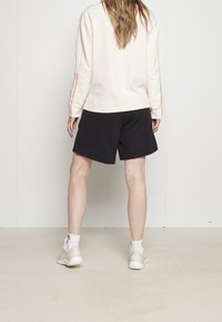 Ljusbeige sweatshirt med röda ränder på ärmarna, i kombination med svarta shorts. Modellen har på sig vita sportskor och ankelsockor.