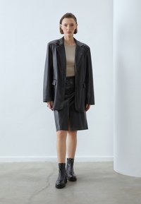 Blazer negro de cuero oversize con falda de cuero a juego que presenta una abertura frontal. Llevado sobre una blusa beige y combinado con botines negros con cremallera hasta el tobillo.
