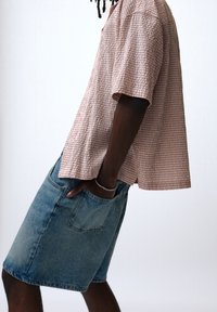 Personne portant une chemise rose à carreaux à manches courtes et une jupe en jean bleue, la main dans la poche, debout sur un fond blanc.