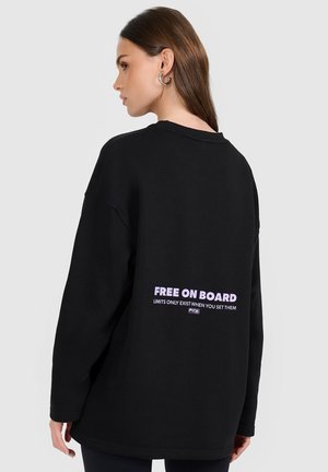 Kobieta z długimi brązowymi włosami, nosząca srebrne kolczyki-koła i czarną bluzę z napisem "Free On Board" na plecach.