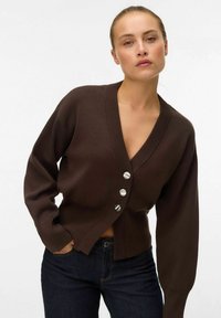 Mujer vestida con un cárdigan marrón oscuro abotonado con tres botones plateados y vaqueros azul oscuro, posando con una mano en el bolsillo.