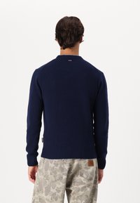 Dunkelblau gestrickter Pullover mit rundem Kragen, gerippten Bündchen und Saum, versehen mit einem kleinen Logo-Detail am Nacken. Kombiniert mit gemusterten beigen Hosen.