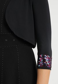 Černý oděv s drapovaným předním dílem a tříčtvrtečními rukávy. Rukáv je zdoben barevnými flitry; tělo je ozdobeno malými cvočky.